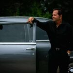 Angelo Oddo, on set of a film, standing next to a car with an open door